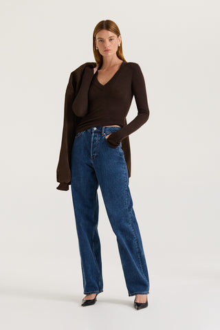 Model wearing a chocolate long-sleeve top and blue jeans, holding a chocolate jacket against a white background.