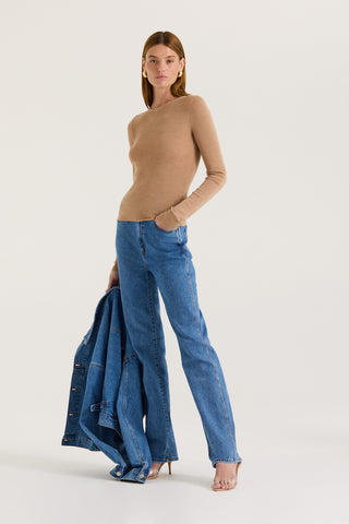 Model wearing a tanned long sleeve top and blue jeans holding a denim jacket with a white background