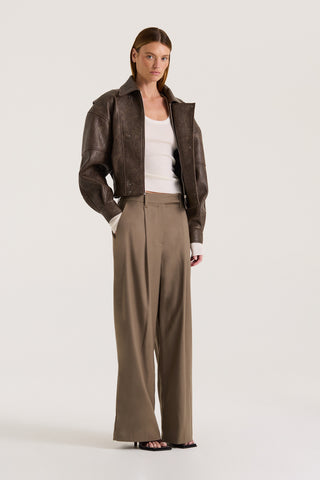 Model wearing a cream long-sleeve scoop neck top, taupe wide-leg pants and a brown jacket on a white background
