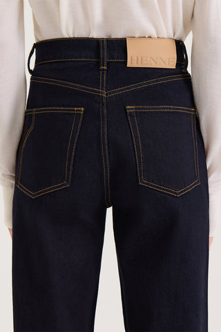 Close up back view of model wearing dark indigo high rise, straight leg jeans with a Henne leather patch on a white background