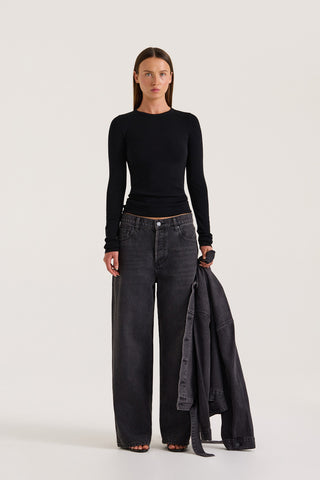Woman wearing a black long-sleeve top and black wide leg jeans holding a black jacket on a white background