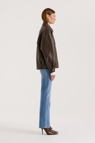 Side view of a model wearing a chocolate leather-effect jacket and blue jeans against a white background.