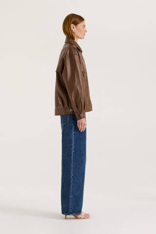 Side view of a model wearing a brown leather effect jacket and blue jeans on a white background