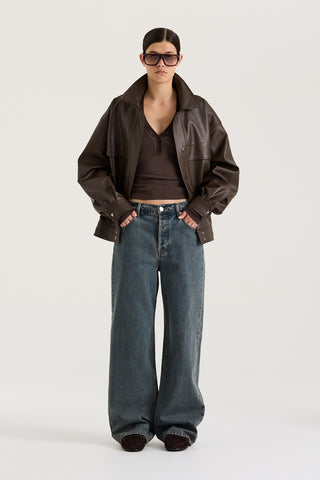 Female model wearing graphite wash wide leg and low rise Henne Gisella Jeans styled with a brown leather jacket on a white background